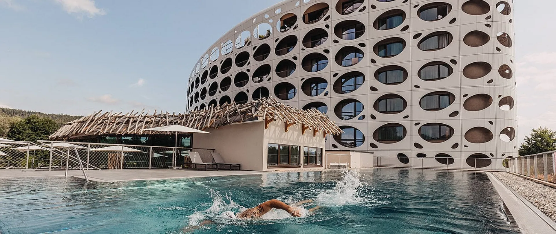 Außenansicht des Das Seepark Hotel mit markanter runder Fassadenarchitektur und Infinity-Außenpool, in dem ein Gast schwimmt – Symbol für aktive Erholung und außergewöhnliche Designhotellerie der Pletzer Resorts.